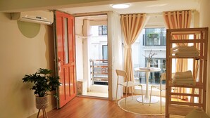 Une pièce avec un plancher en bois, une plante en pot, une porte rouge, une petite table ronde avec une chaise, et une étagère avec des serviettes pliées.