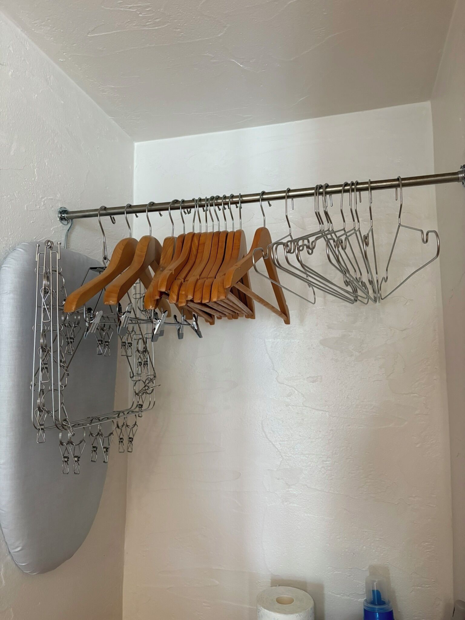 Canto de um quarto com um varal de metal contendo cabides de madeira e prendedores de metal, uma parede branca e uma almofada cinza.