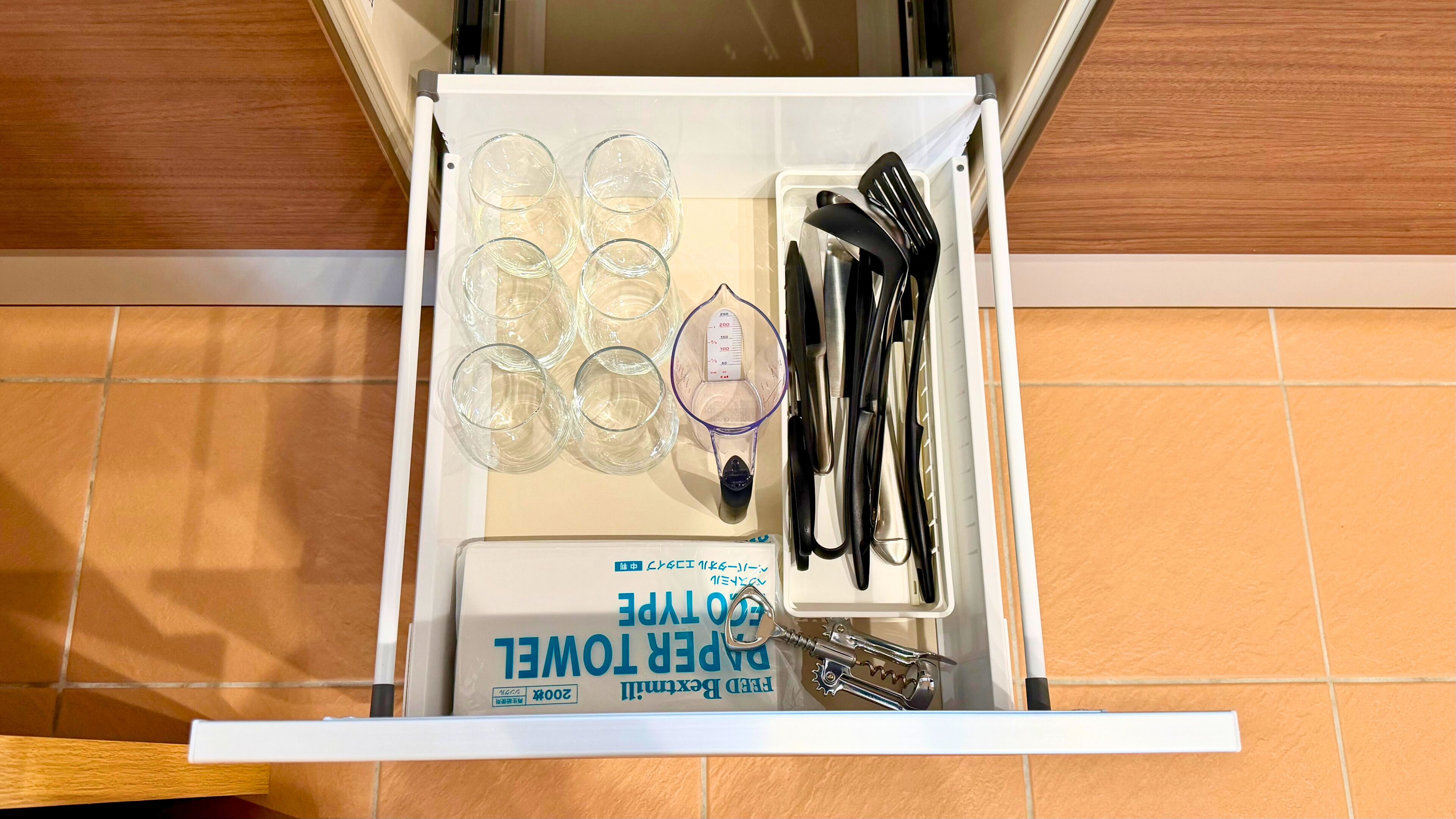 Un cajón de cocina con vasos de vidrio transparente, un vaso medidor, utensilios con mango negro y una caja de toallas de papel.