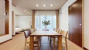 Una habitación amplia con una gran mesa de comedor de madera, sillas color beige, un sofá y un amplio ventanal con cortinas traslúcidas.