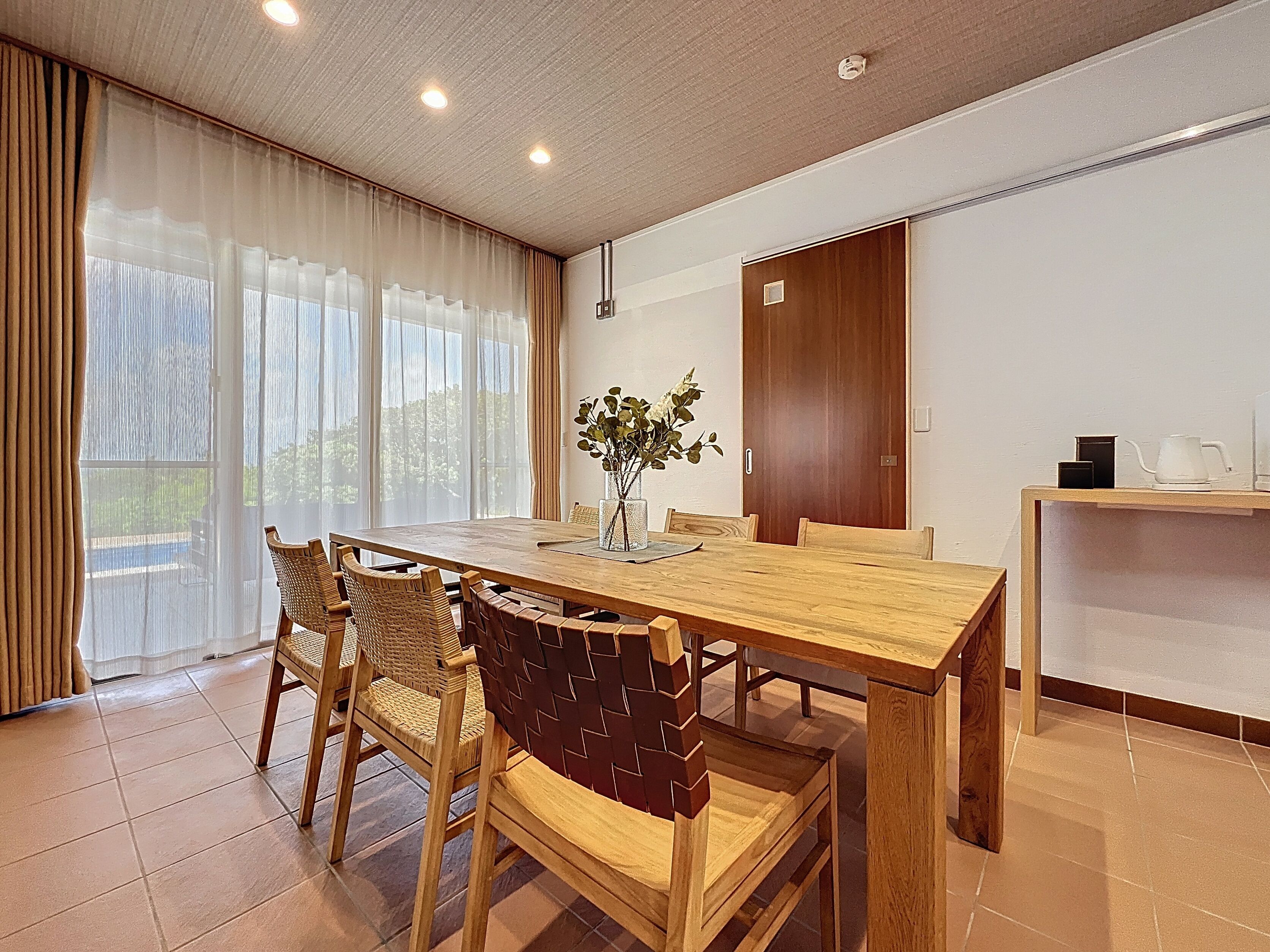 A dining area with a wooden table, wicker chairs, a vase with flowers, and a view of greenery outside.