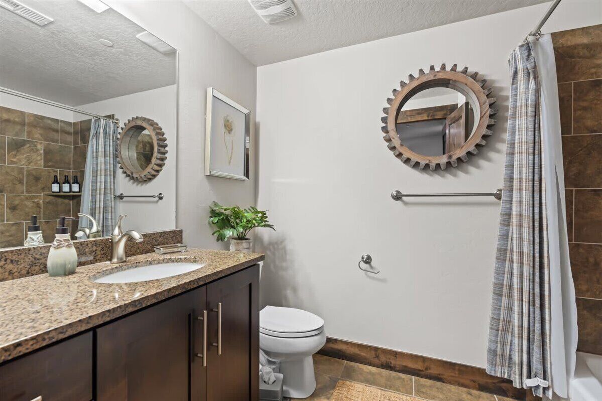 A bathroom with a granite countertop, a white sink, a toilet, and a shower with a curtain. There are two wall-mounted mirrors and a framed picture on the wall.