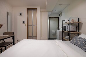 A hotel room with a bed, a desk, a chair, a wardrobe, a shelf with a microwave, and a small bathroom. - Modern Life in Tokyo Nakano 302, High-Rise Residence (都沼袋)