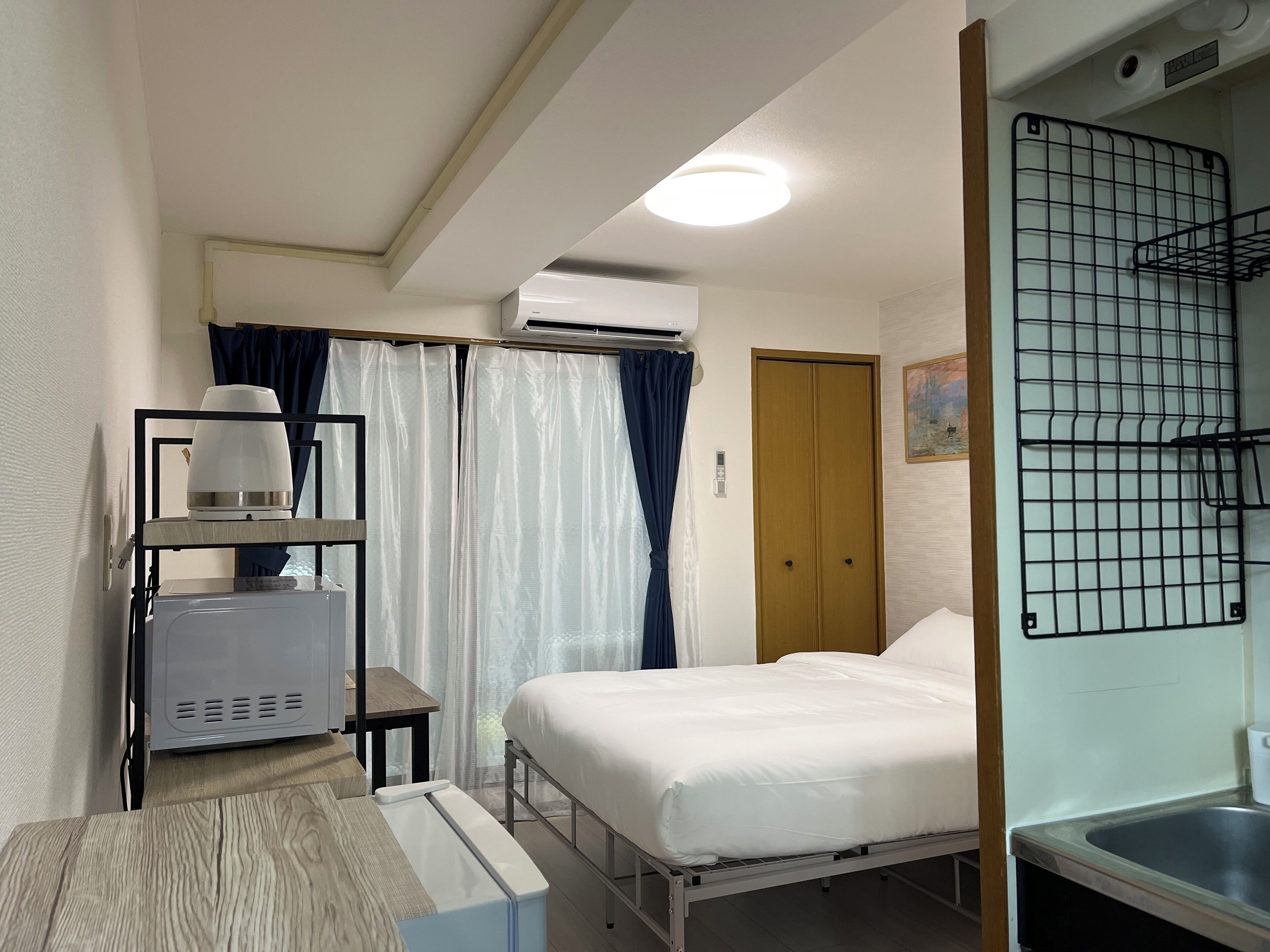 Ein Hotelzimmer mit einem Bett, einem Nachttisch mit Lampe, einer Klimaanlage, einem Fenster mit Vorhängen sowie einer kleinen Küchenzeile mit Spüle.