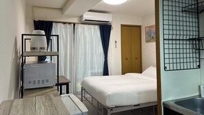 Ett hotellrum med en säng, ett nattduksbord med en lampa, en luftkonditioneringsanläggning, ett fönster med gardiner och ett litet pentry med en diskho.