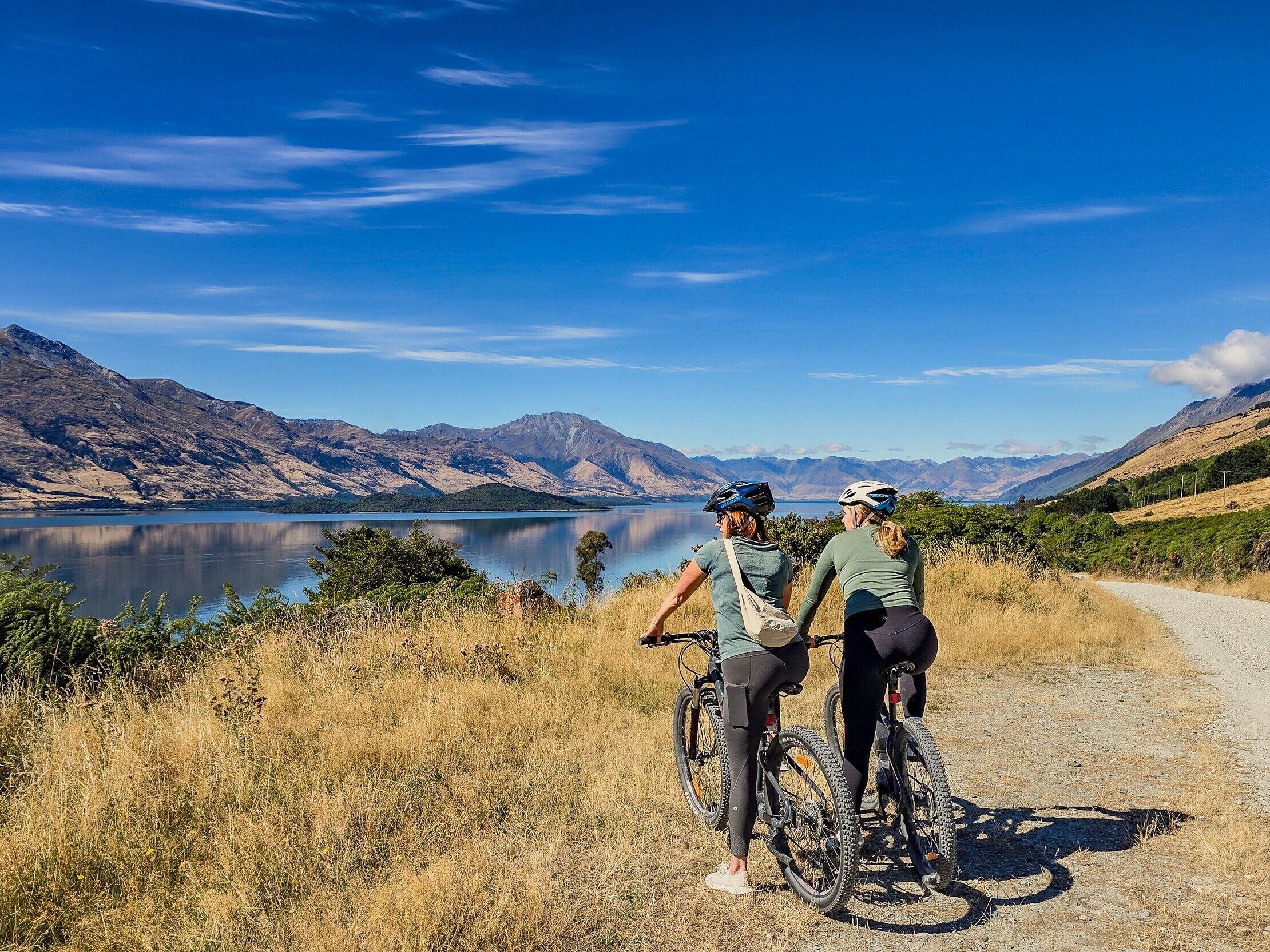 Two cyclists on a gravel path with a lake and mountains in the background.