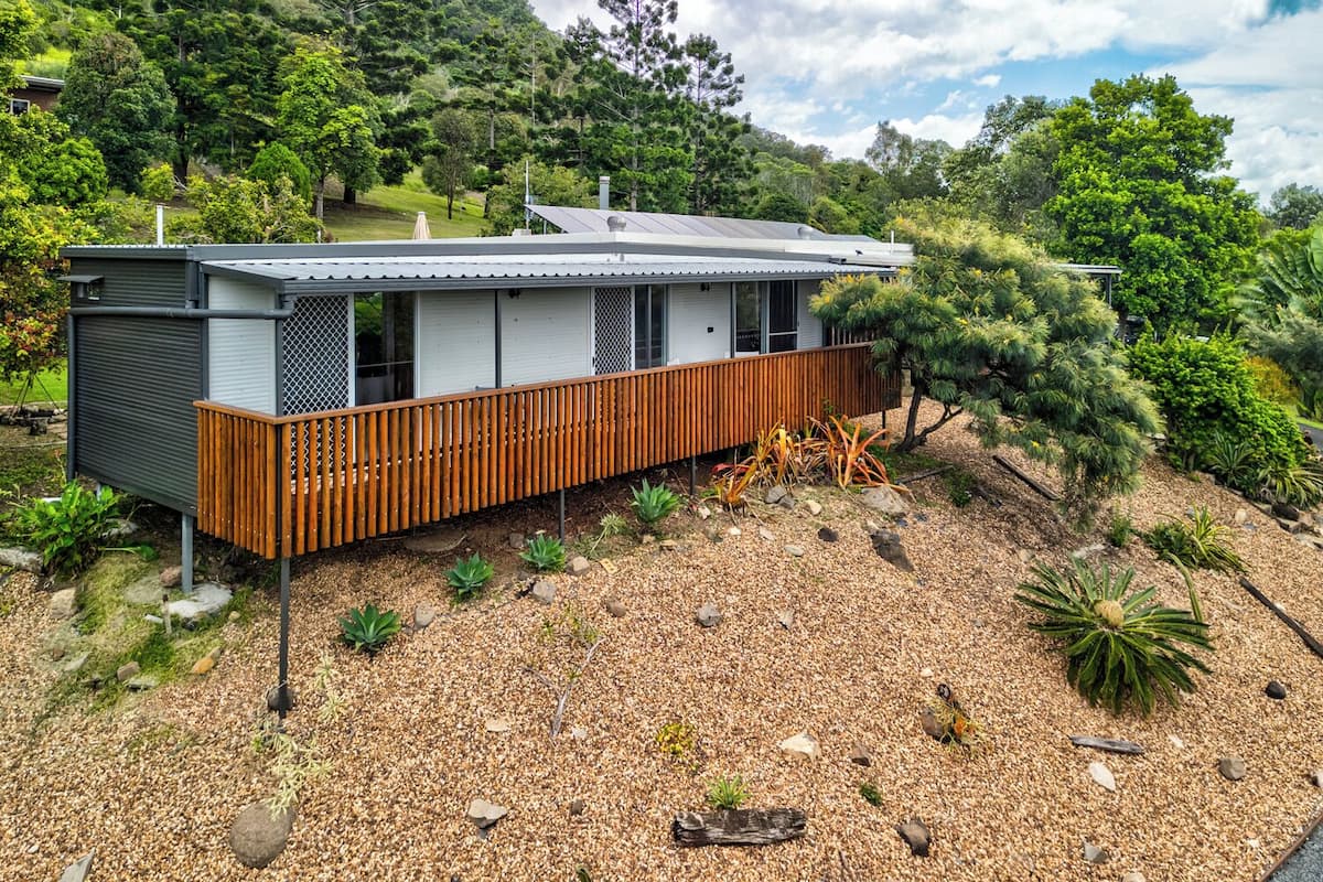 A modern house with a wooden deck, surrounded by a gravel driveway and various plants.