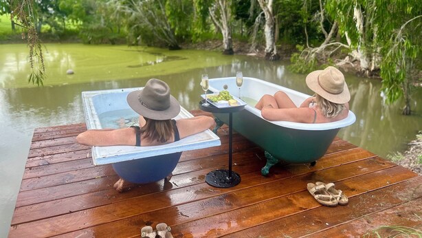 Dos personas relajándose en bañeras sobre un muelle de madera junto a un río tranquilo, con follaje de fondo.