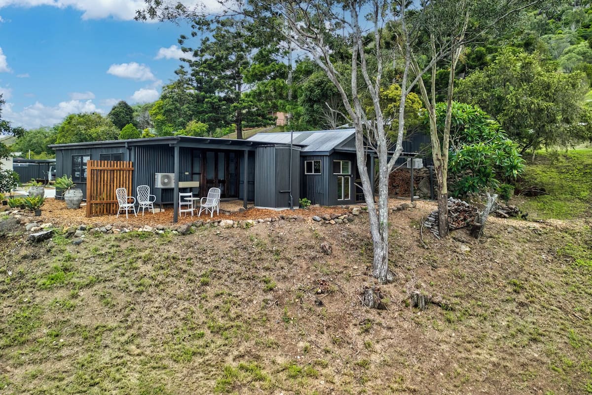 A modern house with a dark exterior, a covered patio with chairs, and a natural surrounding.