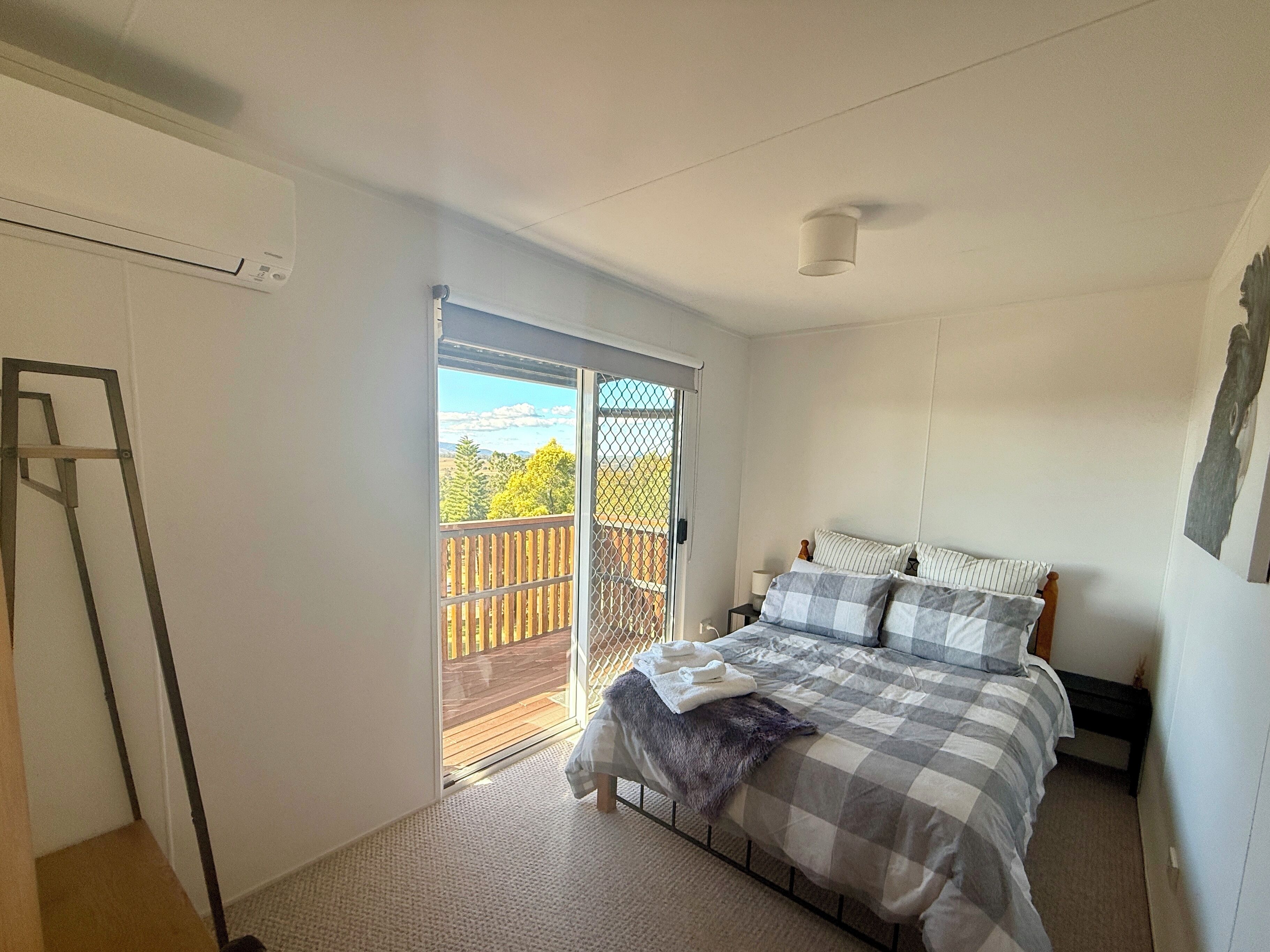 A bedroom with a bed, a small bedside table, a nightstand, a window with a view of a wooden deck and trees, and a sliding glass door leading to an outdoor area.