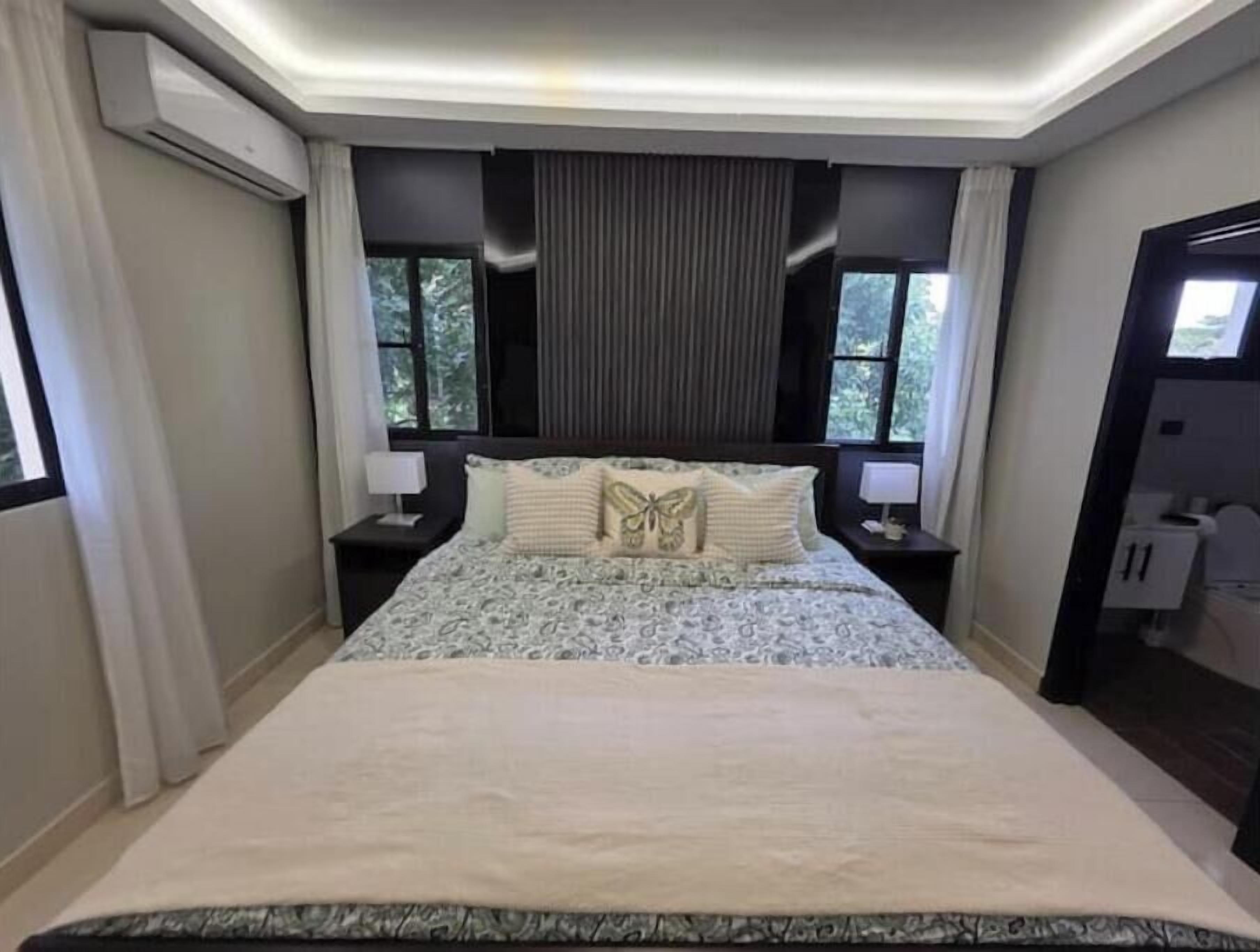 A bedroom with a large bed, two bedside tables with lamps, a window with curtains, and an air conditioning unit.