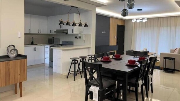 Una cocina moderna con armarios blancos, una zona de comedor con sillas negras y platos rojos, e iluminación colgante.