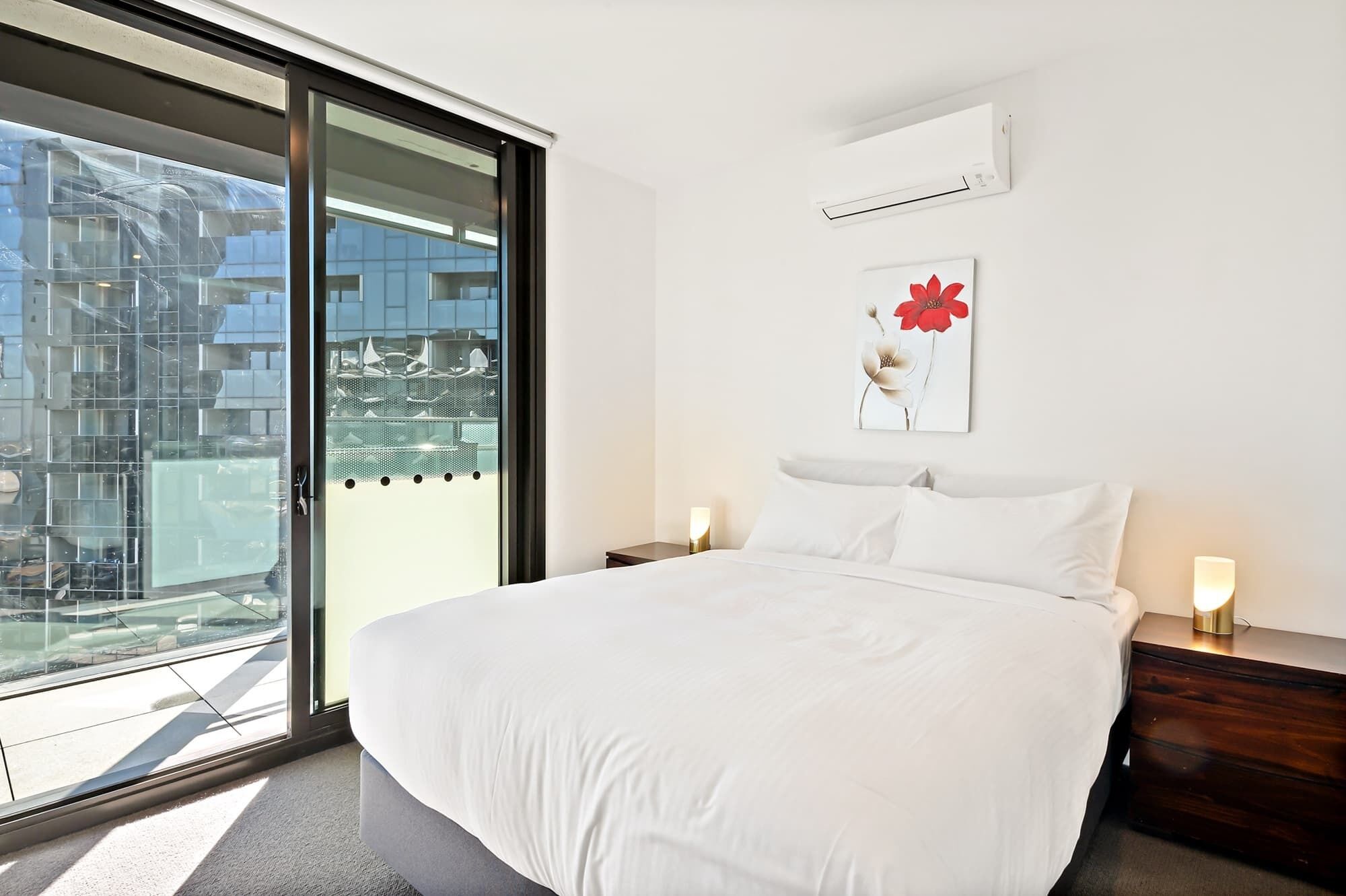 Un dormitorio con una cama grande, una puerta corrediza al balcón, aire acondicionado de pared y una obra de arte enmarcada sobre la cama.