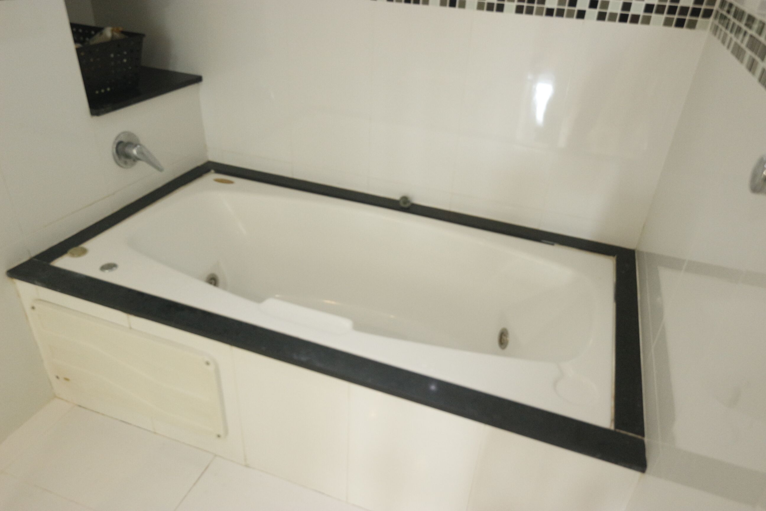 A standard bathtub with a black edge, a chrome faucet, and a white tiled wall with a small shelf above it.