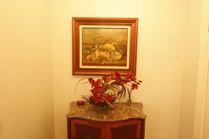 Ein gerahmtes Bild mit Obst und Blumen an der Wand, eine Vase mit roten Blumen auf einem Marmortisch und ein Holzschrank darunter.