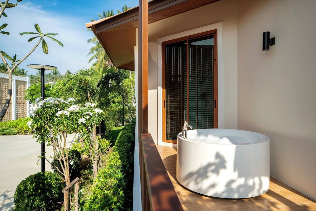 A modern outdoor bathroom with a large bathtub, surrounded by lush greenery and a wooden deck.
