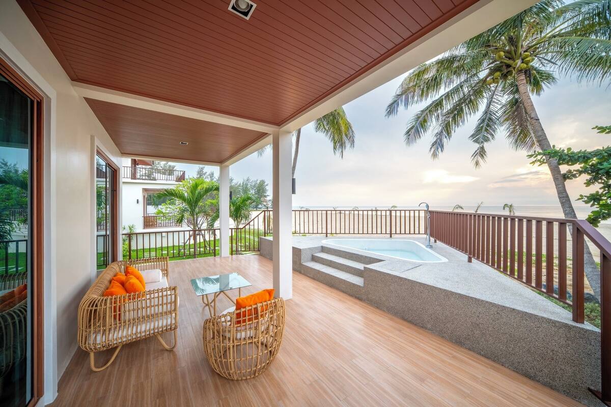 A covered outdoor area with a pool, wooden flooring, wicker furniture, and a view of the sea and palm trees.