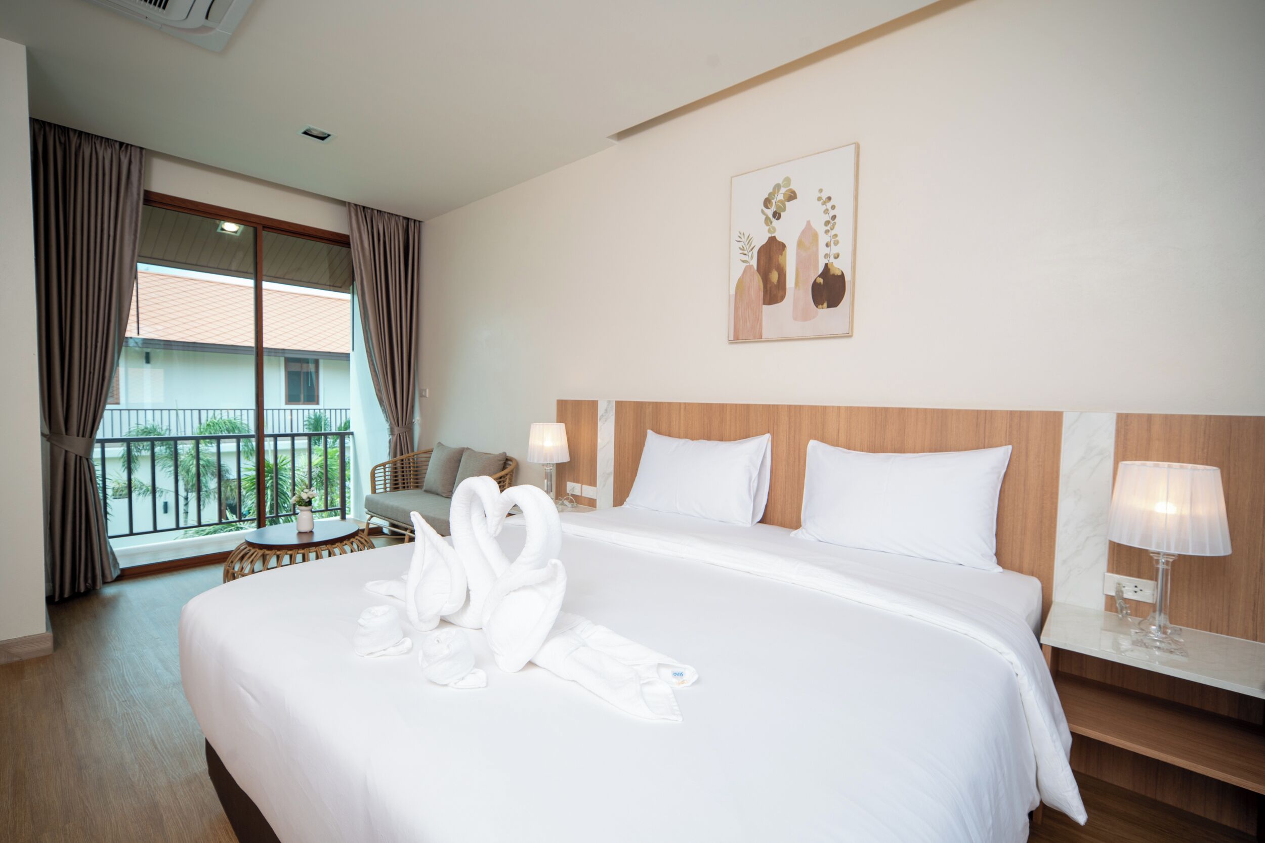 A hotel room with a large bed, a wooden headboard, a balcony with curtains, a small table with a lamp, and a painting on the wall.