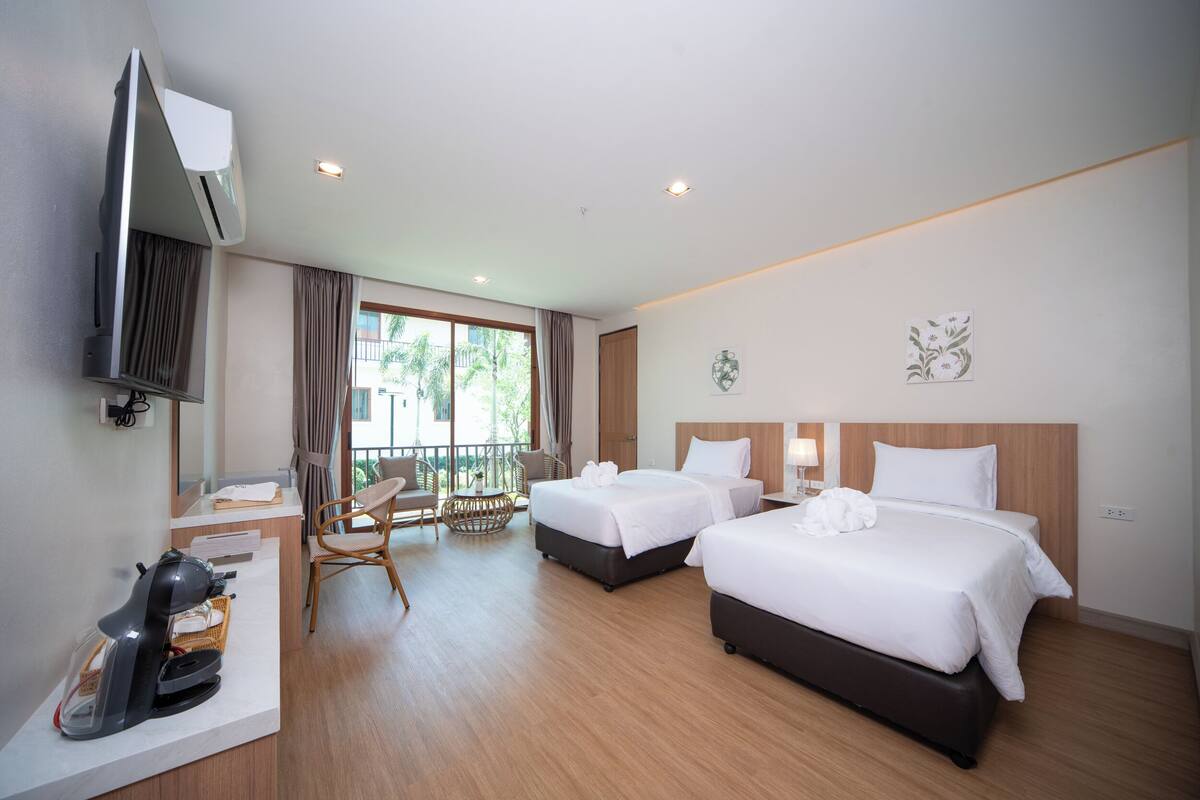 A hotel room with two beds, a TV, a desk with a coffee maker, and a balcony with a view of palm trees.
