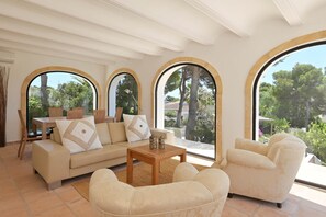 Un salon spacieux avec un canapé beige, des fauteuils et une table basse en bois. De grandes fenêtres en arc offrent une vue sur la verdure et les arbres.