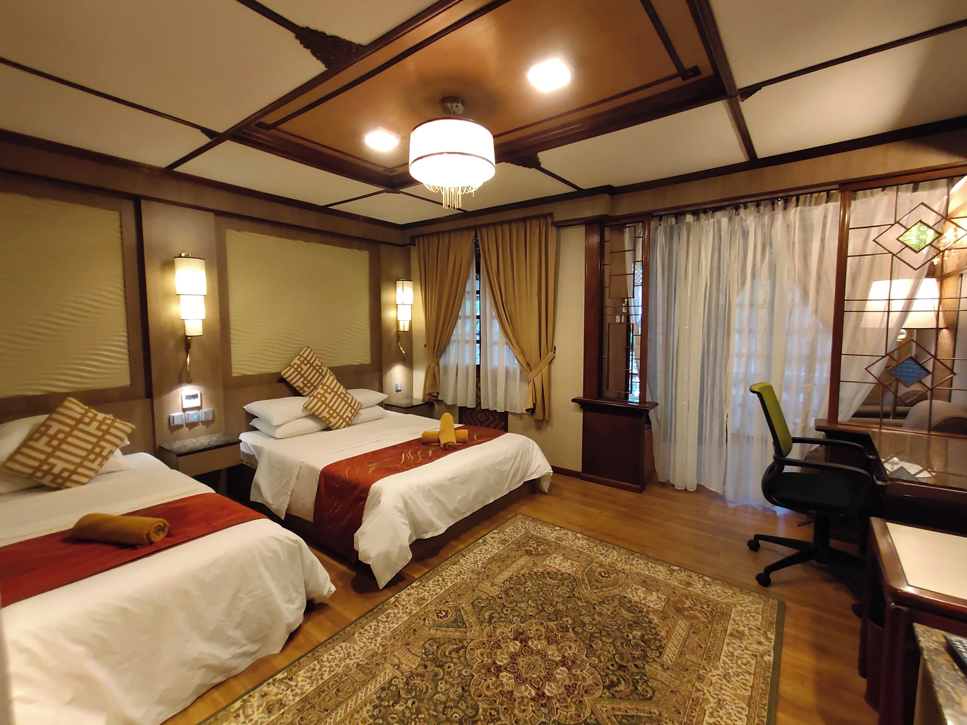 Een hotelkamer met twee bedden, een bureau, een stoel, een groot tapijt en een kroonluchter.