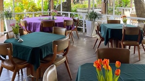 Un restaurante con varias mesas, algunas cubiertas con manteles verdes y morados, y sillas. Hay plantas en macetas y vistas de follaje al exterior.