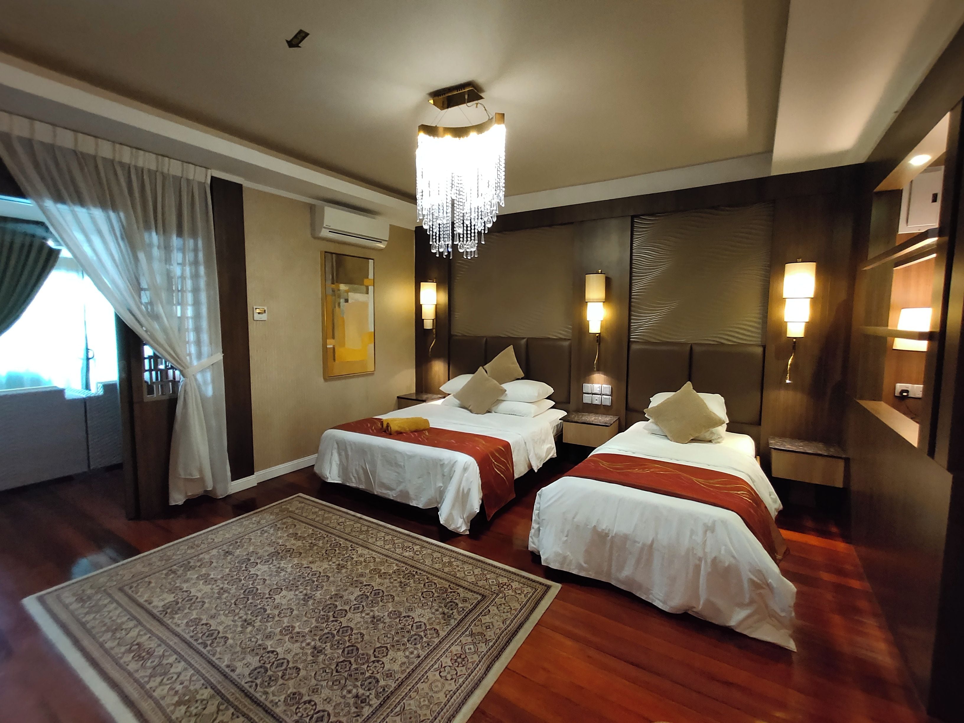 A hotel room with two beds, a large rug, a chandelier, and a view of the outdoors.
