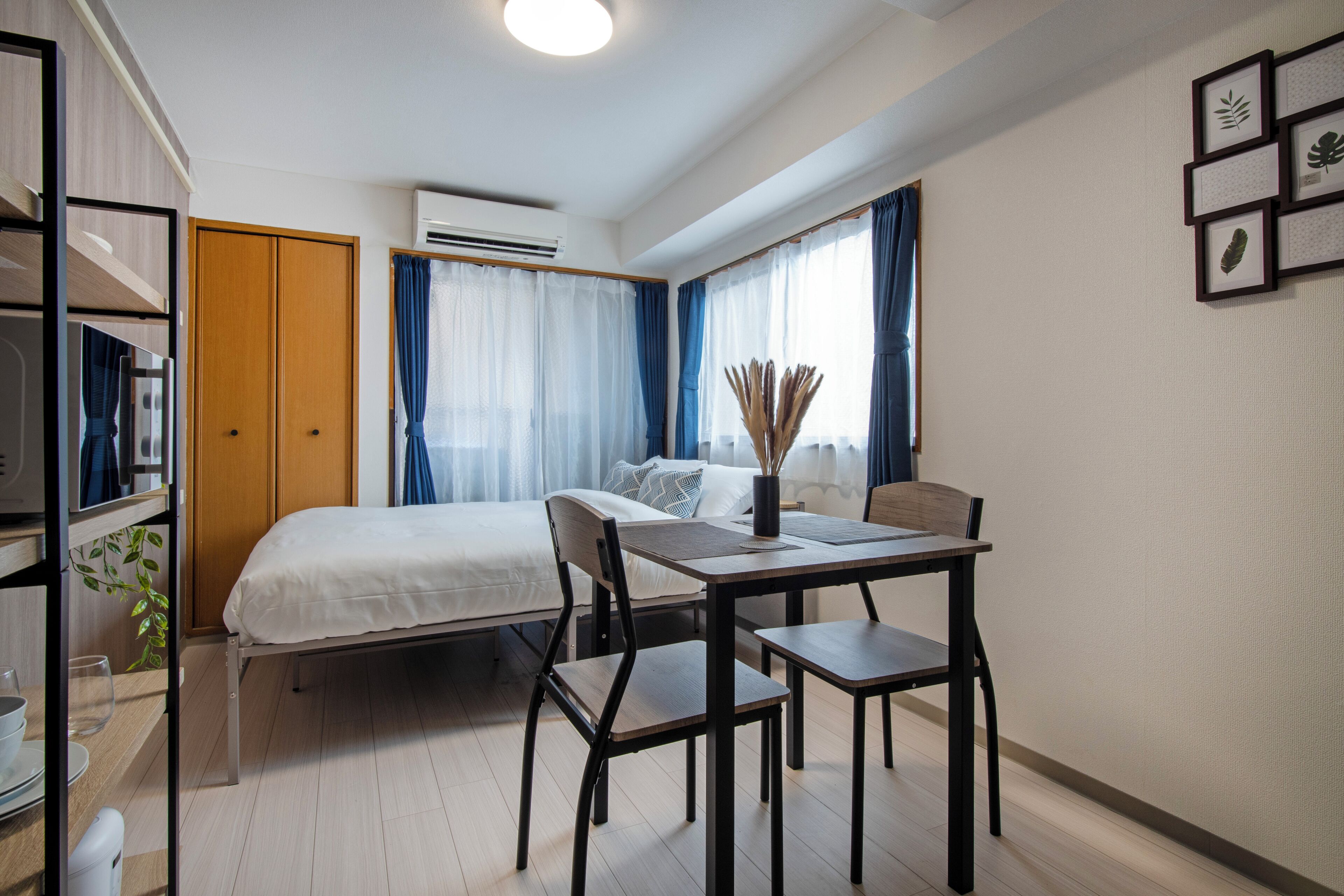 Et lille hotelværelse med seng, spisebord med stole, et tekøkken og et vindue med gardiner.
