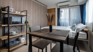 Una camera d'albergo compatta con un letto, una scrivania con una sedia, un forno a microonde, scaffali con stoviglie e una finestra con tende.