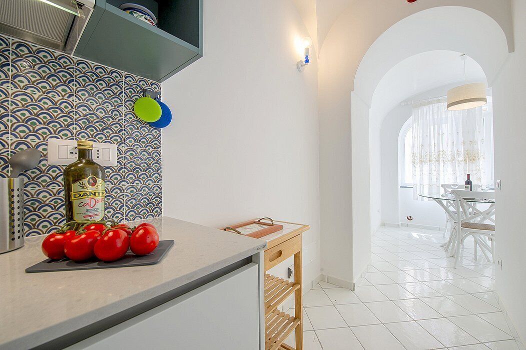 Uma cozinha com painel de azulejos, bancada com tomates, uma garrafa de azeite e uma mesa lateral de madeira.