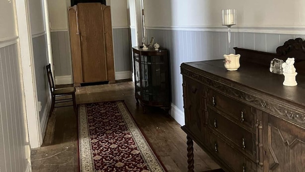 A hallway with a wooden door, a dark wooden dresser, a red patterned rug, and a white wall with decorative molding. - Lovely 4-bedroom villa, save on kennel fees and bring your dog! (Waihi)