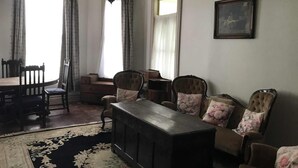 A living room with a dark wooden TV stand, floral-patterned chairs, a dining table with chairs, and a large window with curtains. - Lovely 4-bedroom villa, save on kennel fees and bring your dog! (Waihi)