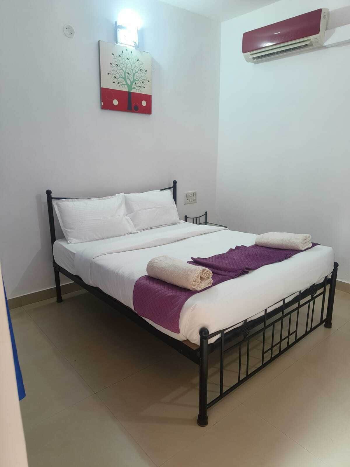 Una cama individual con ropa blanca y una manta morada, un aire acondicionado de pared y un adorno de pared con un árbol.