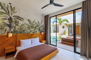 A modern hotel room with a large bed, wooden headboard, and a view of a swimming pool and outdoor seating area. - Villa Linaria Pecatu (Ungasan)