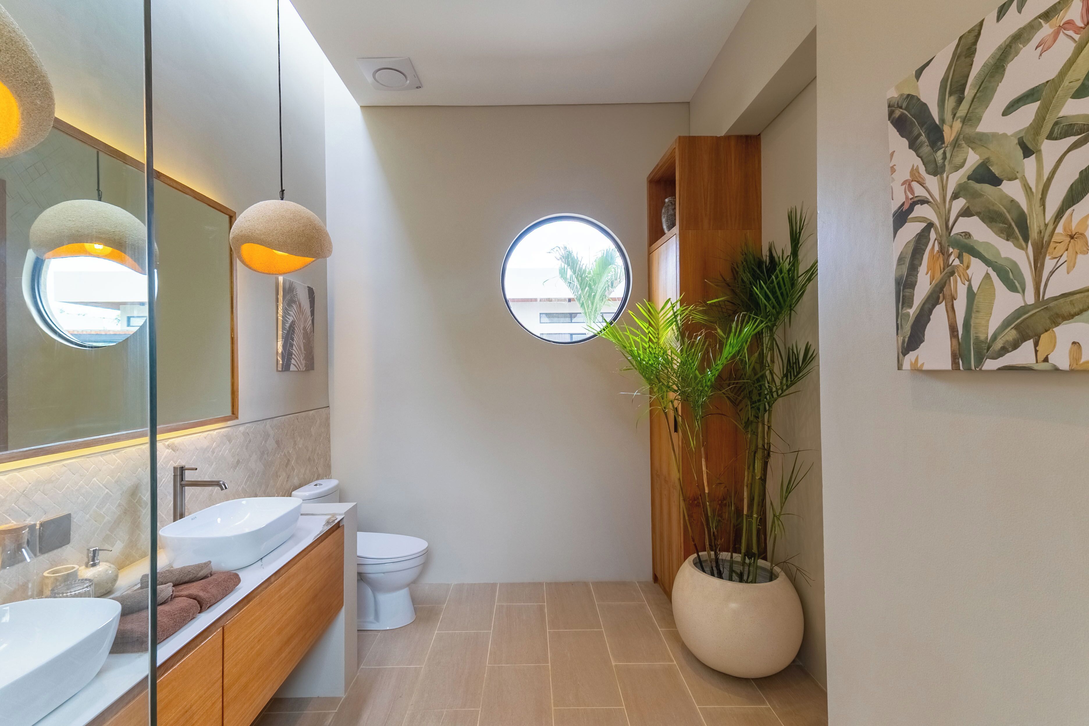 A modern bathroom with a large mirror, two pendant lights, a round window, a toilet, a sink, a vanity, and a potted plant.