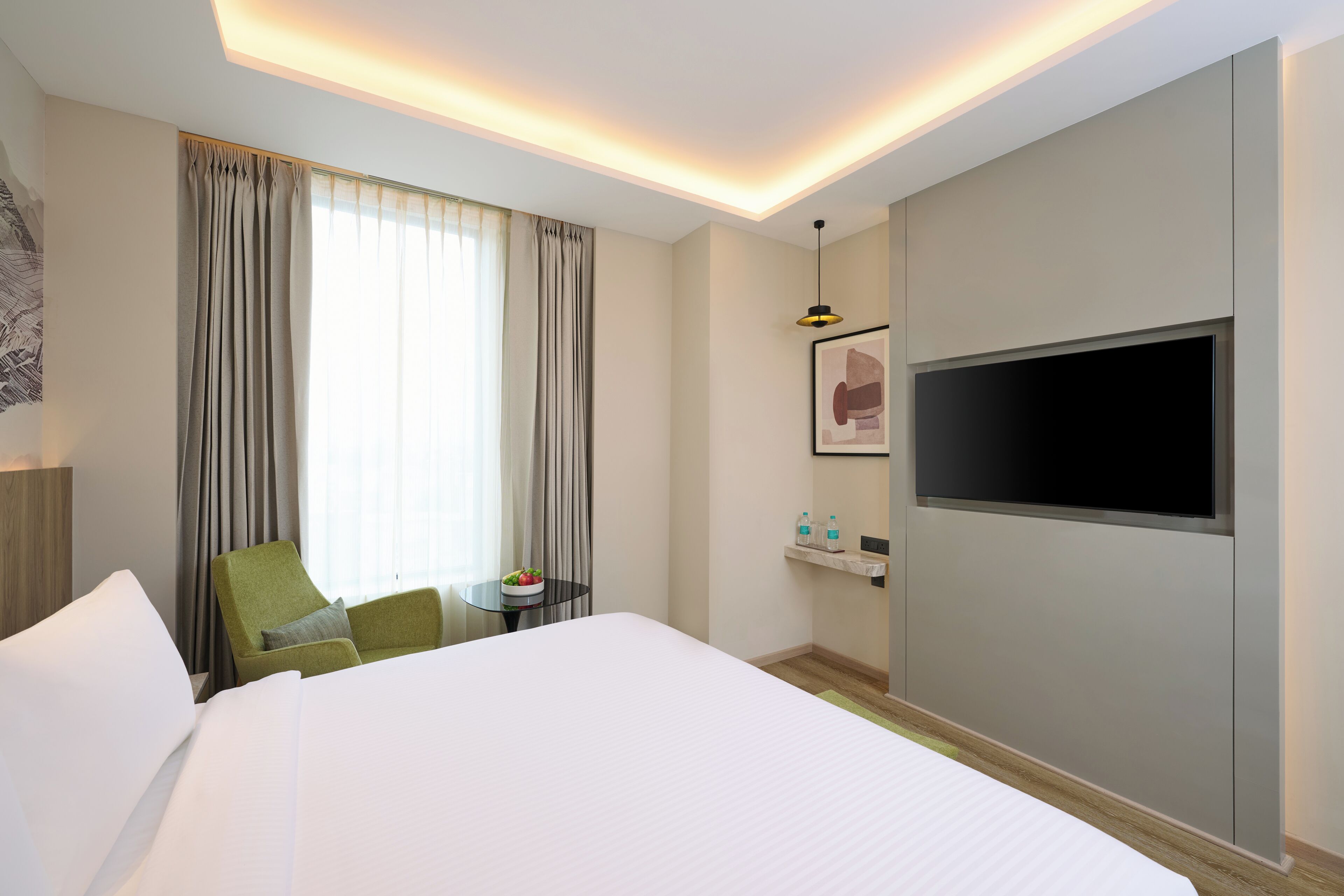 A hotel room with a large bed, a flat-screen TV mounted on the wall, a small table with a bowl of fruit, and a green chair.
