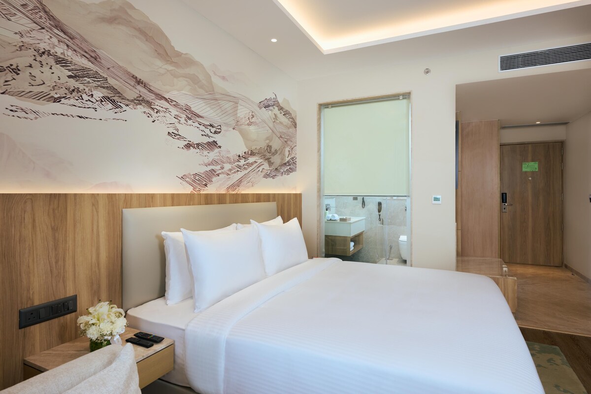 A modern hotel room with a large bed, a nightstand with a remote control, a vase of flowers, and a large wall mural.