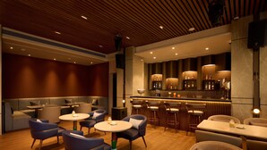 Un área de bar moderna con mesas redondas y taburetes, un espacio de estar con sofás y sillas, y un techo de madera con iluminación empotrada.