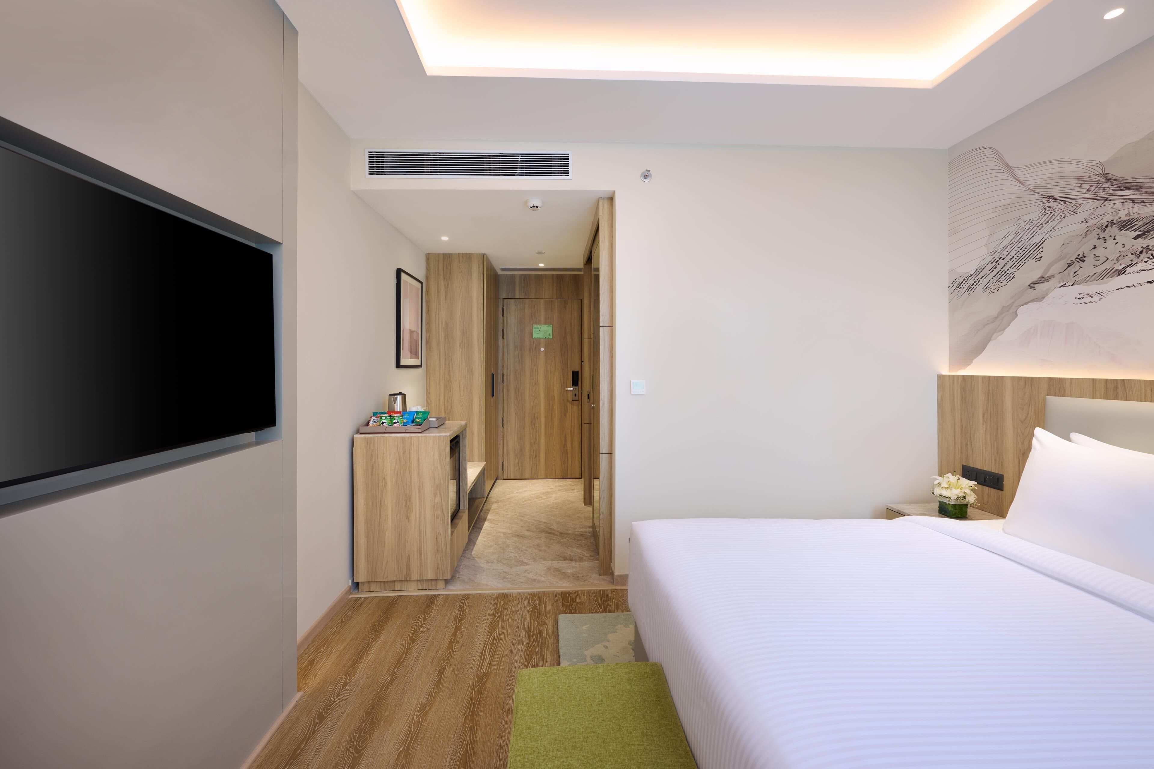 A modern hotel room with a large bed, a flat-screen TV mounted on the wall, a wooden desk with a chair, and a small bathroom area.