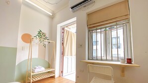 Une pièce avec un lit, un plancher en bois, une fenêtre avec un rideau, une plante suspendue et une chaise blanche.