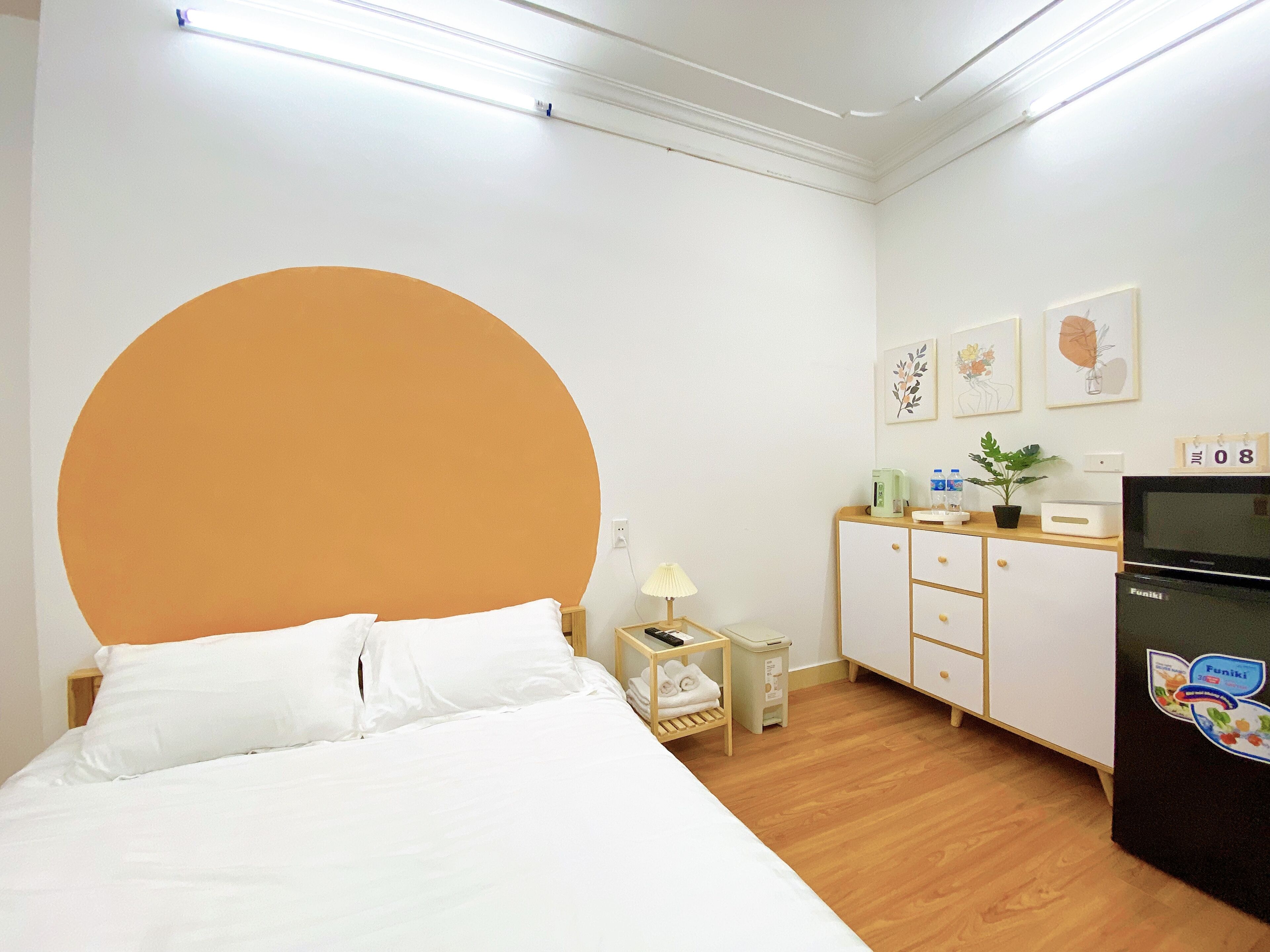 A bedroom with a bed, a dresser, a nightstand, a lamp, a small table with a plant, and a refrigerator.