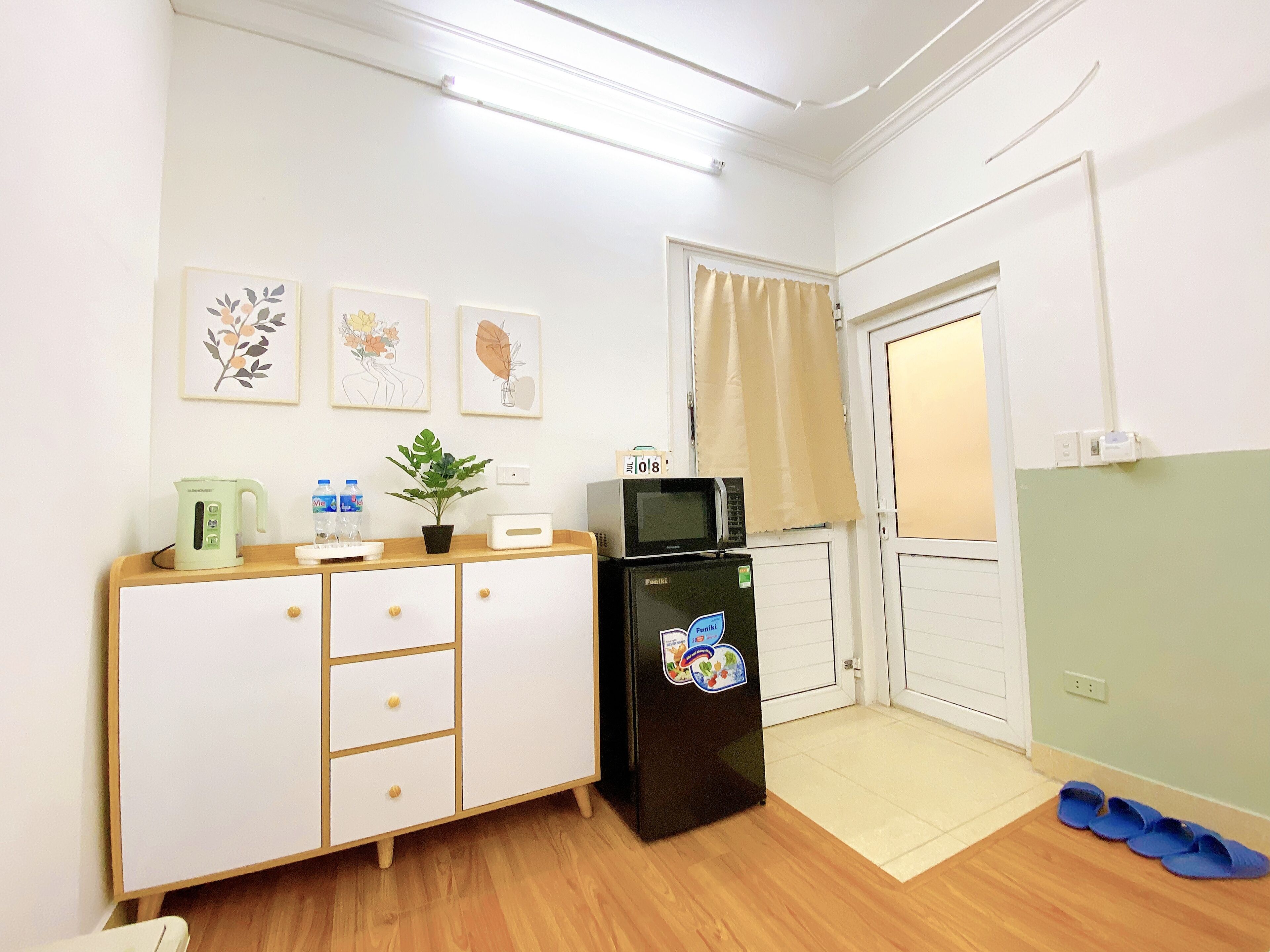 A compact kitchenette with a microwave, kettle, and a small refrigerator. There are framed pictures on the wall and a pair of slippers on the floor.