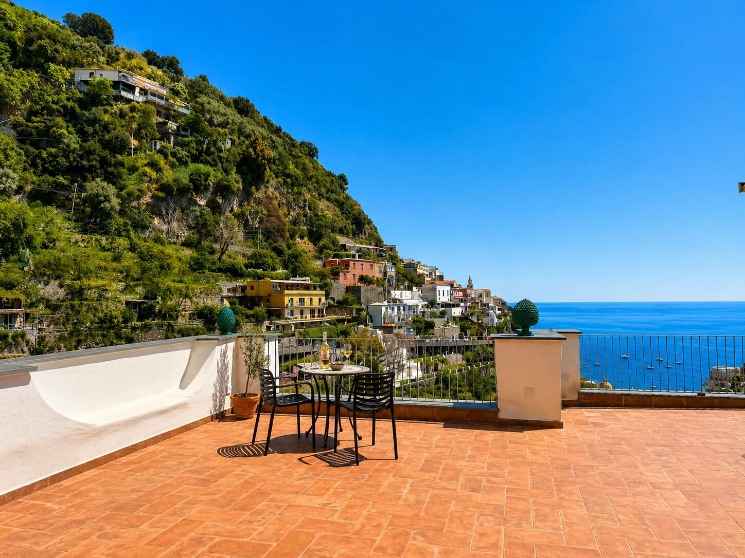 A terrace with a table and chairs, overlooking a coastal town with buildings on a hillside.
