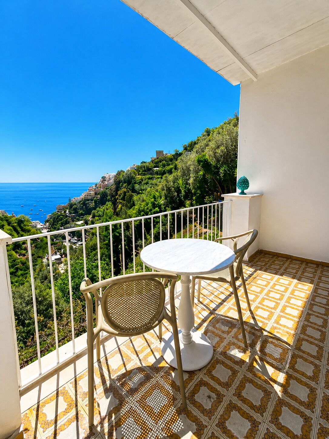 Ein Balkon mit einem weißen Tisch und zwei Stühlen, mit Blick auf die Küste, Gebäude und Grünflächen.
