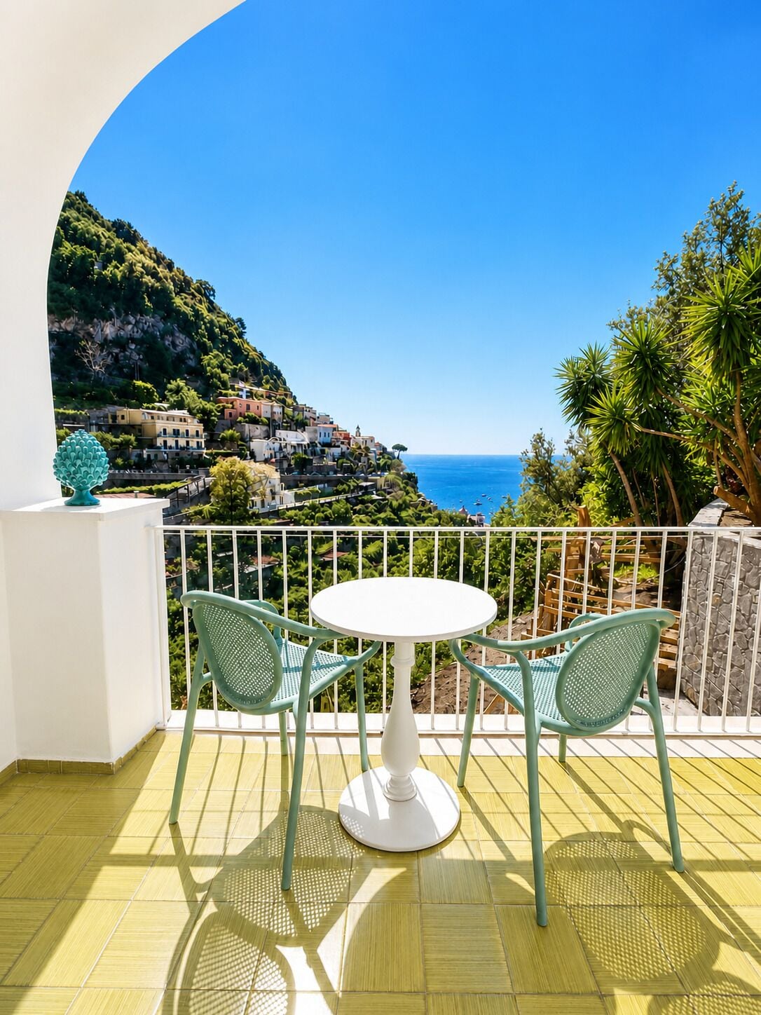 A balcony with a white table and two chairs, overlooking a coastal village with buildings on a hillside.