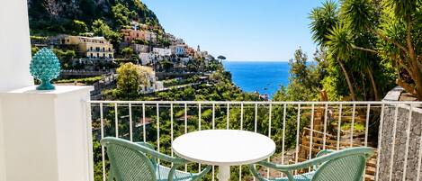 A balcony with a white table and two chairs, overlooking a coastal village with buildings on a hillside.