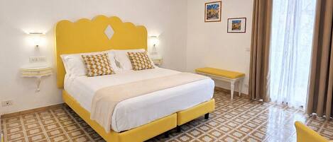 Una camera d'albergo con un letto giallo, piastrelle a pavimento decorate, un comodino, una sedia e una finestra con tende.