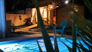 Une piscine éclairée la nuit, avec un espace patio, des palmiers et un bâtiment dont les lumières diffusent une ambiance chaleureuse.