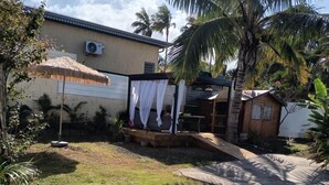 Un jardin avec un parasol en chaume, un espace salon abrité et une petite construction en bois.