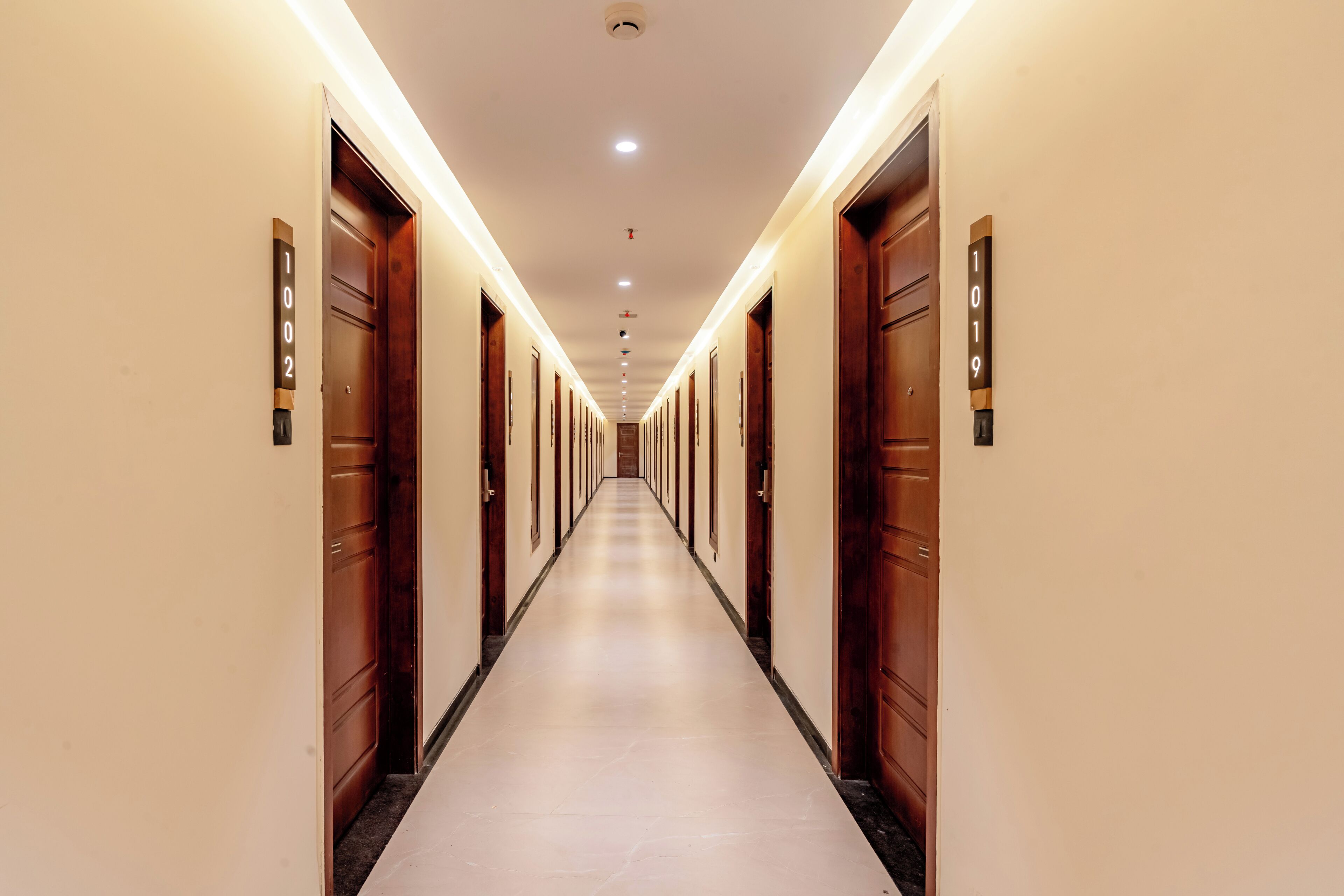 A long, narrow hotel corridor with wooden doors on either side, numbered room signs, and a tiled floor.